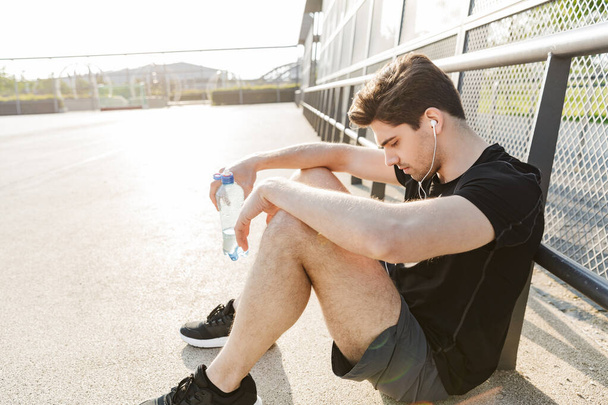 Portrait of brunette man wearing earphones holding water bottle  - Foto, Bild