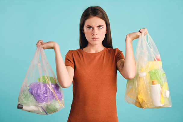 Krásná dívka v tričku drží eko tašky s plastovým odpadem a hněvivě dívá do kamery přes barevné pozadí - Fotografie, Obrázek