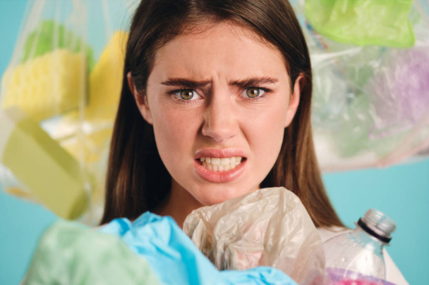 Close up irritated girl with plastic waste around scared looking in camera over colorful background - Photo, Image