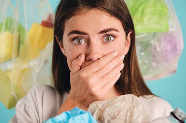 Portrait of shocked girl with plastic waste around amazedly looking in camera over colorful background - Photo, Image