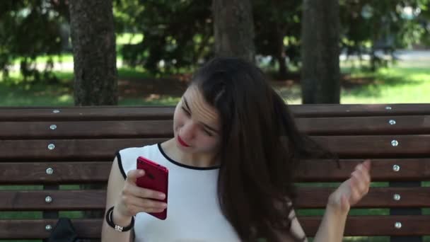Close up portrait A beautiful young girl in a white T-shirt is chatting in social networks on her smartphone while sitting on a bench in a park.A beautiful young girl straightens her hair and posts in social networks on her smartphone. - Video