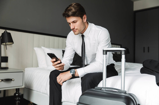 Image of modern young man holding cellphone while sitting on bed - Foto, Bild