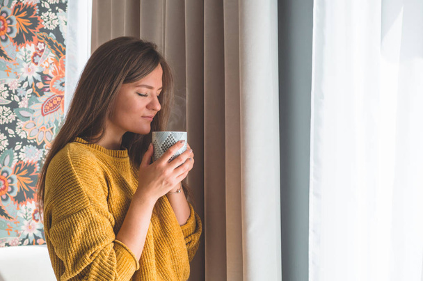 Kobieta z filiżanką gorącego napoju przy oknie. Patrząc przez okno i pijąc herbatę. - Zdjęcie, obraz