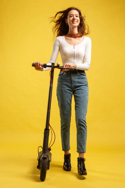 Young beautiful girl with electric scooter. Isolated on yellow background. Technology and freedom of movement. - Фото, зображення