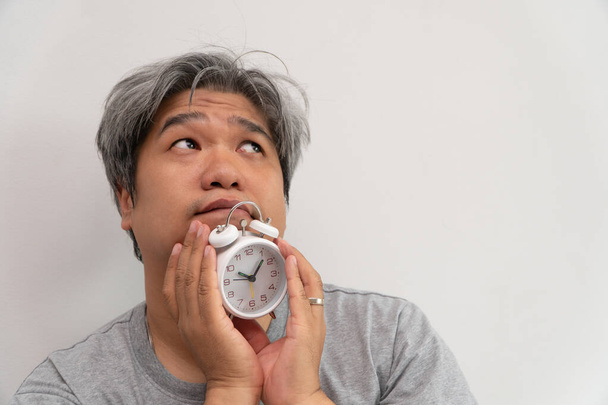 Asyalı orta yaşlı bir adam elinde beyaz bir çalar saat tutuyor. Yüzünde can sıkıntısı var ve kendini kötü hissediyor. Problemi uyku bozukluğu. Uykusuzluk konsepti - Fotoğraf, Görsel
