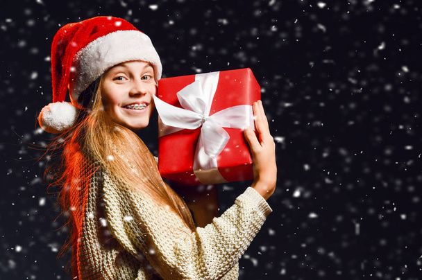 Teenager girl with braces holding hugging her new year gift red box with ribbon at her cheek happy to get under the snow - Fotó, kép