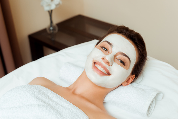 vista de ángulo alto de la mujer sonriente con máscara en la cara acostada en la mesa de masaje en el spa
  - Foto, Imagen