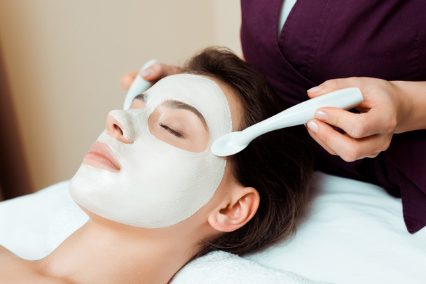 cropped view of cosmetologist applying face mask on attractive woman in spa  - Foto, afbeelding