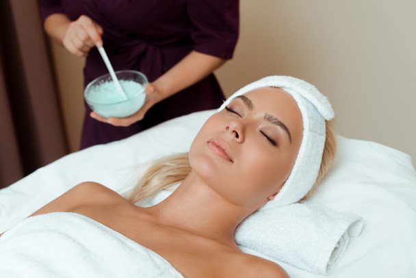 Ausgeschnittene Ansicht des Kosmetikers mit Schale mit Gesichtsmaske und attraktiver Frau im Spa liegend  - Foto, Bild