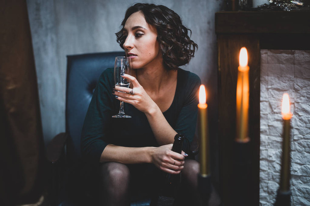 Young beautiful woman in a green dress with a glass of sparkling wine sits in a chair by the fireplace - Фото, изображение