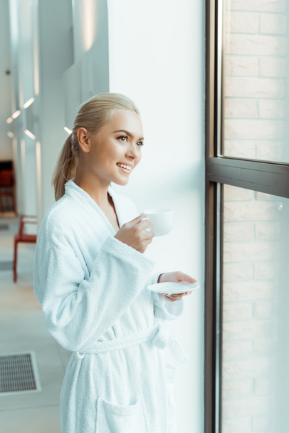 attractive and smiling woman in white bathrobe holding cup of coffee and looking through window in spa  - Foto, Imagem