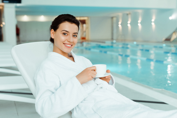 attractive and smiling woman in white bathrobe holding cup of coffee in spa  - Foto, afbeelding