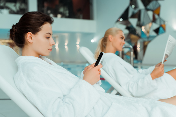 foco seletivo da mulher atraente usando smartphone e amigo ler jornal em segundo plano no spa
  - Foto, Imagem