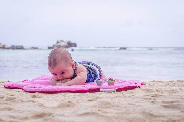 Дитина на пляжі лежить на піску рожевий килимок
 - Фото, зображення