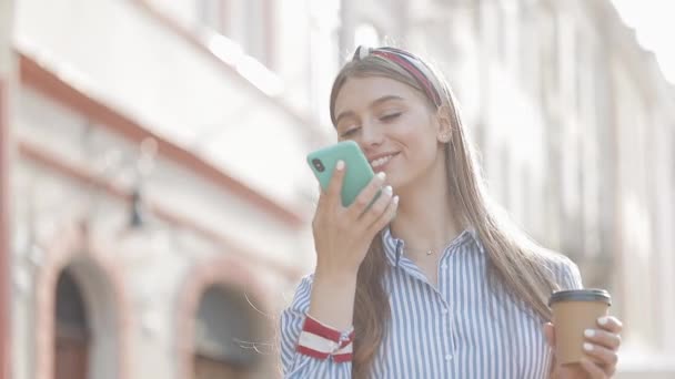 Young Beautiful Caucasian Girl with Brown Hair and Headband on it Wearing Striped Shirt Using her Smartphone. Woman Sending Voice Message and Scrolling Standing at the City Street. - Video, Çekim