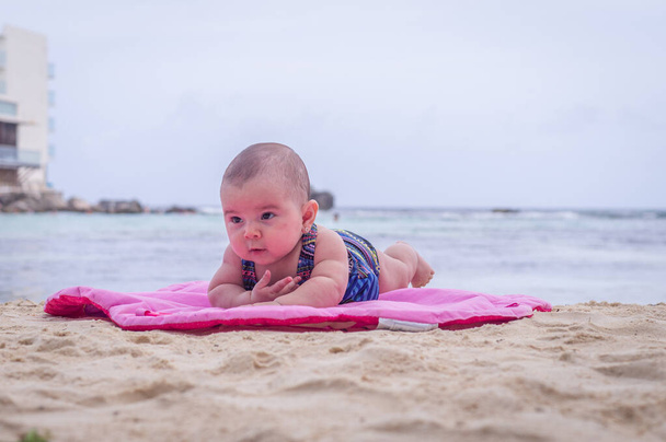 Ребенок на пляже, лежащий на песчано-розовом коврике
 - Фото, изображение