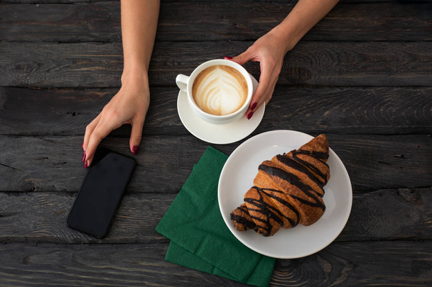 La donna tiene il telefono in una mano e la tazza di cappuccino in un'altra. Croissant su un tavolo di legno scuro. - Foto, immagini