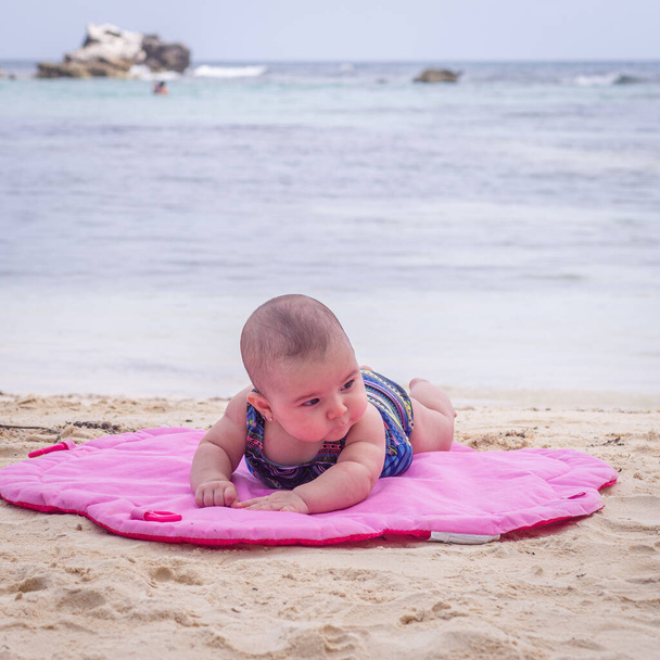 Bebê na praia deitado no tapete rosa areia
 - Foto, Imagem