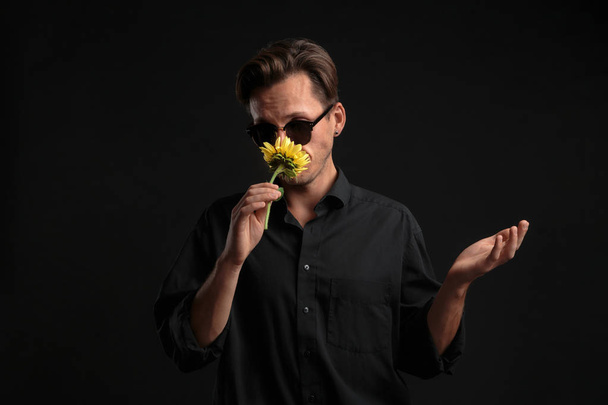 Siyah gömlekli ve güneş gözlüklü seksi adam. Sevdiğine vermek için elinde güzel sarı bir çiçek tutuyor. - Fotoğraf, Görsel