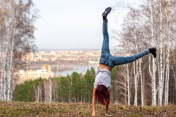 A girl with red hair makes an acrobatic wheel on nature against the background of a blurred city and forest. The girl has red hair, jeans. Motion blur. Grass on the ground. Acrobatics in nature. - Photo, Image