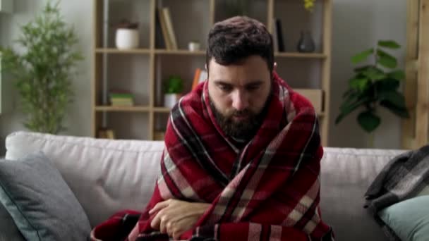 Ill man with chills and wraps himself in a blanket on the sofa in living room - Záběry, video