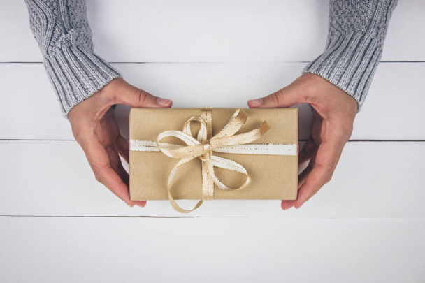 Manos de mujer con suéter gris sosteniendo un regalo con un lazo dorado sobre fondo de madera blanca. Navidad, año nuevo, cumpleaños, día de San Valentín, concepto del día de la madre. Vista superior
. - Foto, imagen