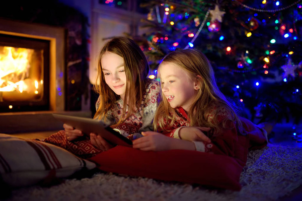 Two cute young sisters using a tablet pc at home by a fireplace in warm and cozy living room on Christmas eve. - Foto, Imagem