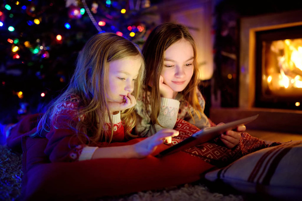 Two cute young sisters using a tablet pc at home by a fireplace in warm and cozy living room on Christmas eve. - Foto, Bild