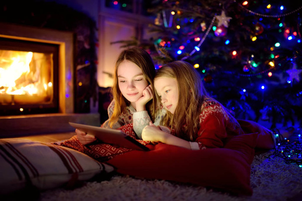 Two cute young sisters using a tablet pc at home by a fireplace in warm and cozy living room on Christmas eve. - Φωτογραφία, εικόνα