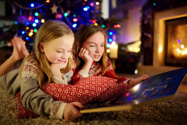 Happy young sisters reading a story book together by a fireplace in a cozy dark living room on Christmas eve. Celebrating Xmas at home. - Фото, изображение
