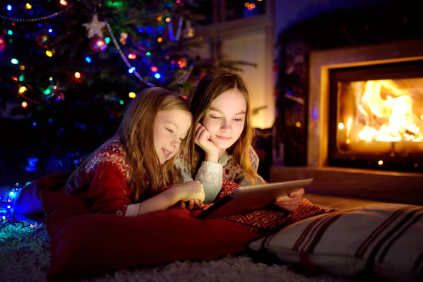 Two cute young sisters using a tablet pc at home by a fireplace in warm and cozy living room on Christmas eve. - Fotoğraf, Görsel