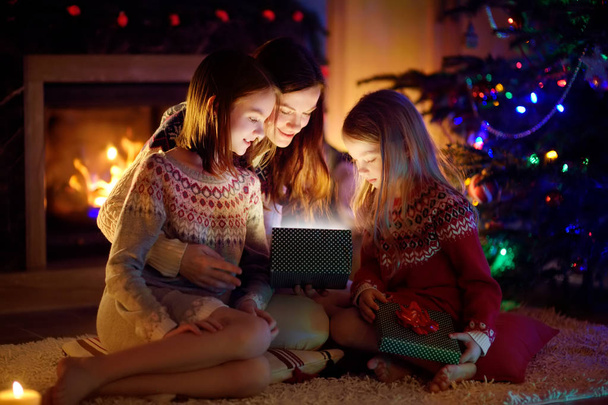 Happy young mother and her two small daughters opening a magical Christmas gift by a fireplace in a cozy dark living room on Christmas eve. - Fotó, kép