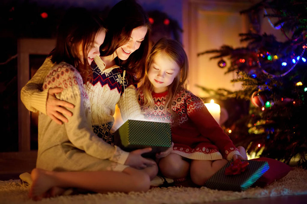 Happy young mother and her two small daughters opening a magical Christmas gift by a fireplace in a cozy dark living room on Christmas eve. - Фото, зображення