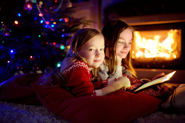Two cute young sisters using a tablet pc at home by a fireplace in warm and cozy living room on Christmas eve. - Foto, Imagem