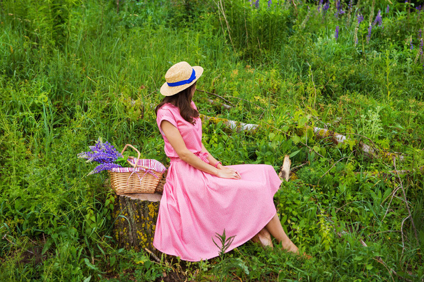 piękna dziewczyna w pięknej sukience siedzi na pniu w lesie, bukiet łubinu w koszyku - Zdjęcie, obraz