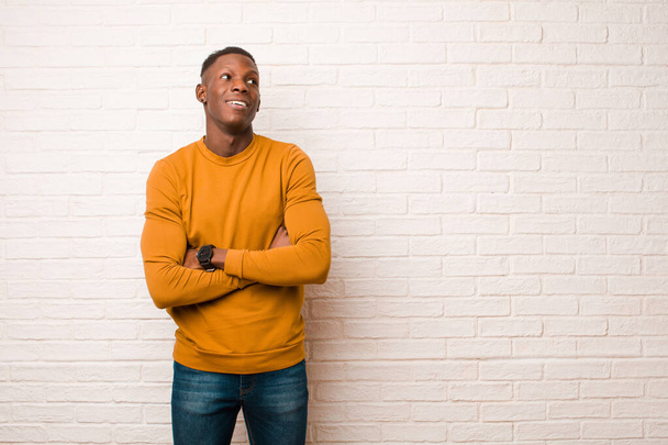 young african american black man feeling happy, proud and hopeful, wondering or thinking, looking up to copy space with crossed arms against brick wall - Фото, изображение