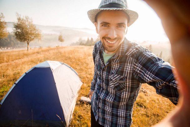 Φάνυ γενειοφόρος άντρας χαμογελάει και βγάζει σέλφι στα βουνά από το κινητό του. Ταξιδιώτης με γενειάδα φορώντας καπέλο να λάβει αυτο πορτρέτο με κάμερα μετά την πεζοπορία φθινόπωρο. - Φωτογραφία, εικόνα