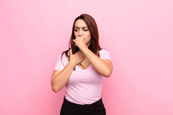 young pretty woman feeling ill with a sore throat and flu symptoms, coughing with mouth covered against pink wall - Photo, image