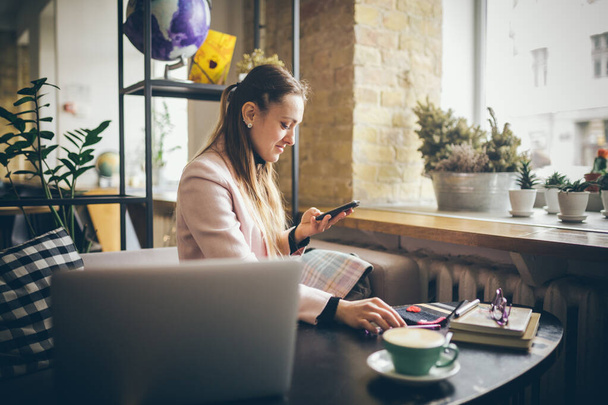 nainen istuu kahvilassa puinen pöytä, kahvin juominen ja älypuhelimen. Nainen käyttää puhelinta kahvilassa. Opiskelija opiskelee verkossa. Tyttö tarkistaa sähköpostin. Nainen Cafe, käytä matkapuhelinta, Working Notebook
 - Valokuva, kuva