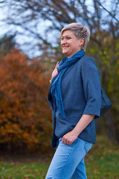 Onnellinen vaalea nainen kävelee syksyn kaupungin puistossa, pukeutunut sinisiin farkkuihin ja takkiin
 - Valokuva, kuva