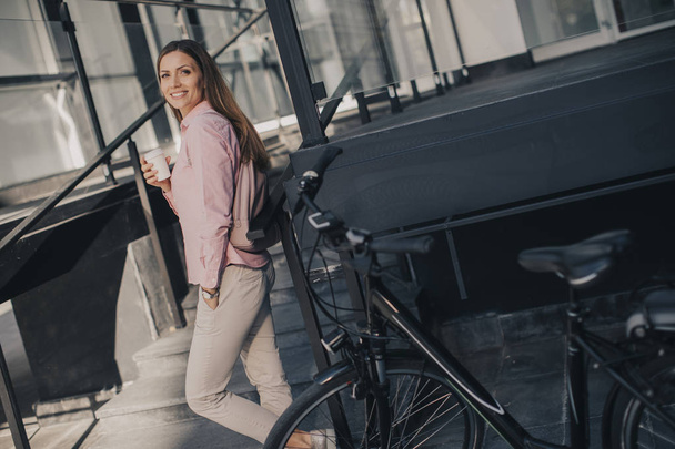 Kaunis nuori naispyöräilijä juo kahvia sähköpolkupyörän kupista kaupunkiympäristössä
 - Valokuva, kuva