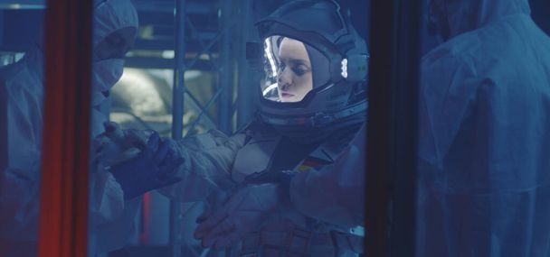 Tiedemies auttaa astronauttia laittamaan hanskat käteen.
 - Valokuva, kuva