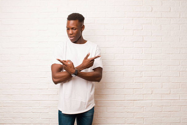 young african american black man looking puzzled and confused, insecure and pointing in opposite directions with doubts against brick wall - Foto, Bild