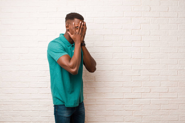 young african american black man feeling scared or embarrassed, peeking or spying with eyes half-covered with hands against brick wall - Foto, imagen