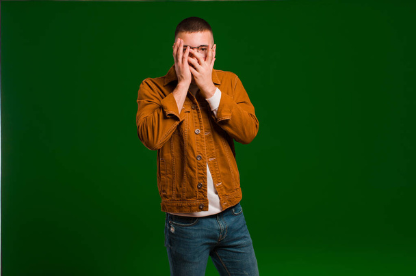 young handsome man feeling scared or embarrassed, peeking or spying with eyes half-covered with hands against flat background - Фото, изображение
