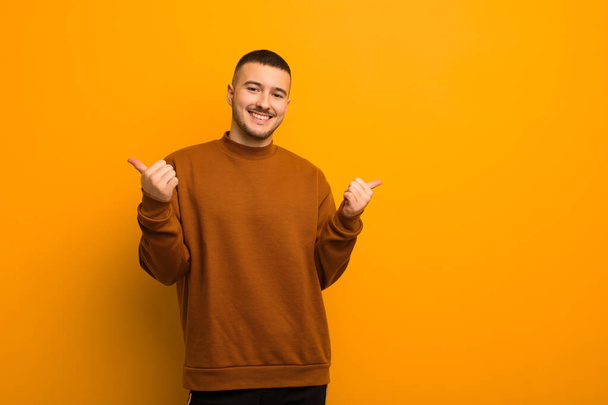 young handsome man smiling joyfully and looking happy, feeling carefree and positive with both thumbs up against flat background - Foto, Imagen