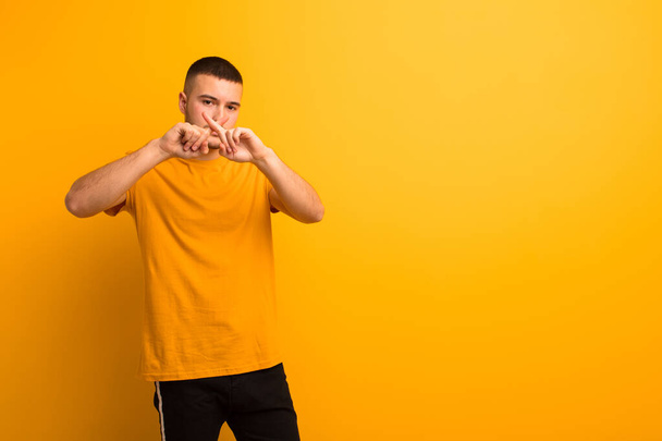 young handsome man looking serious and displeased with both fingers crossed up front in rejection, asking for silence against flat background - Foto, Imagen