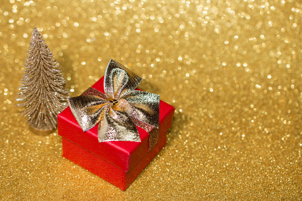Piros ajándék doboz arany íj és kis karácsonyfa arany csillogó felületen bokeh fények.  - Fotó, kép