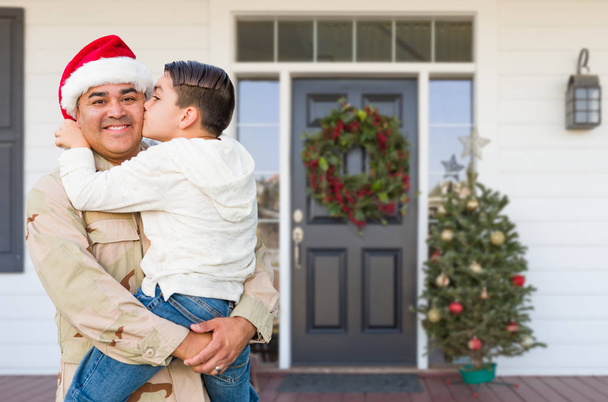 hispanische Soldat mit Weihnachtsmütze hält Sohn gemischter Rasse vor Haus - Foto, Bild