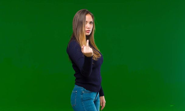 young pretty woman feeling angry, annoyed, rebellious and aggressive, flipping the middle finger, fighting back against green background - Foto, Bild